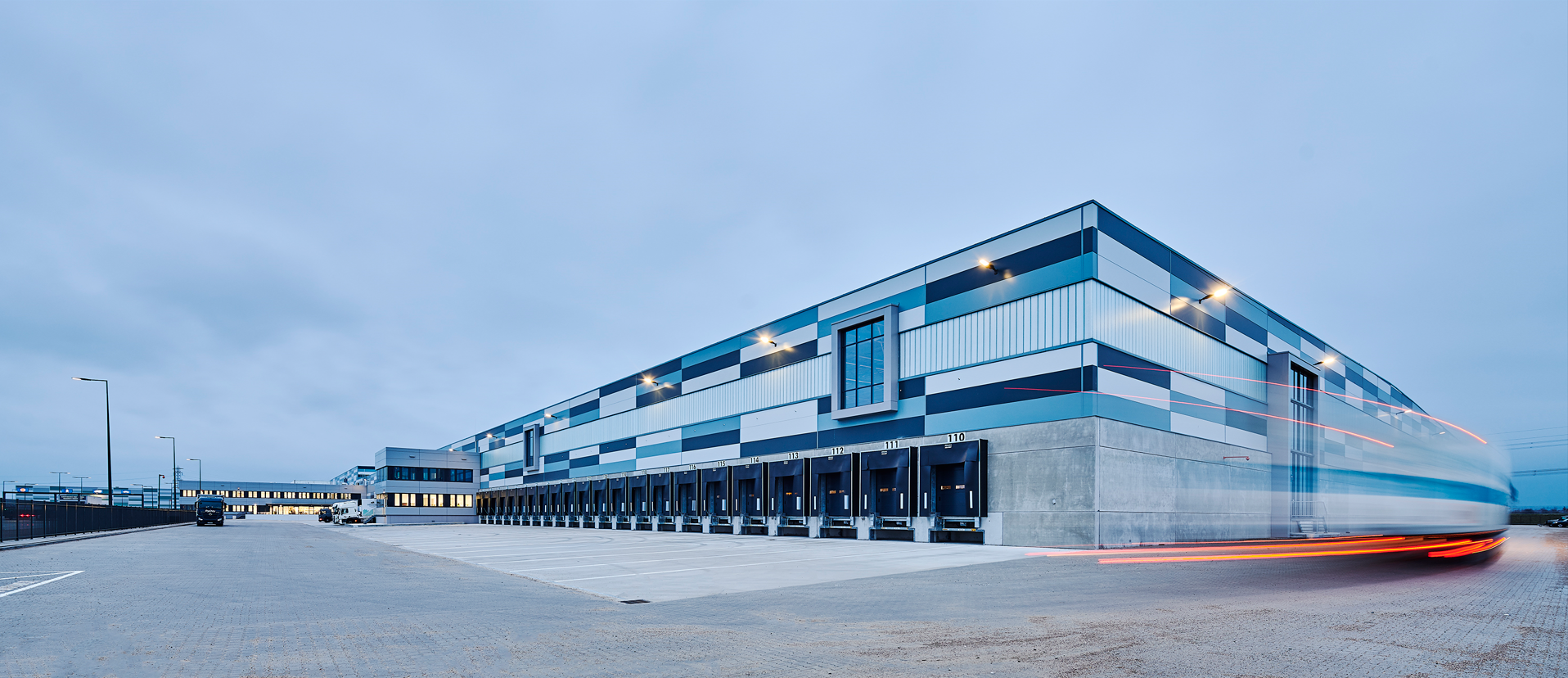 Zgrada logističkog centra u zoru, zablurano vozilo koje brzo prolazi pored zgrade.